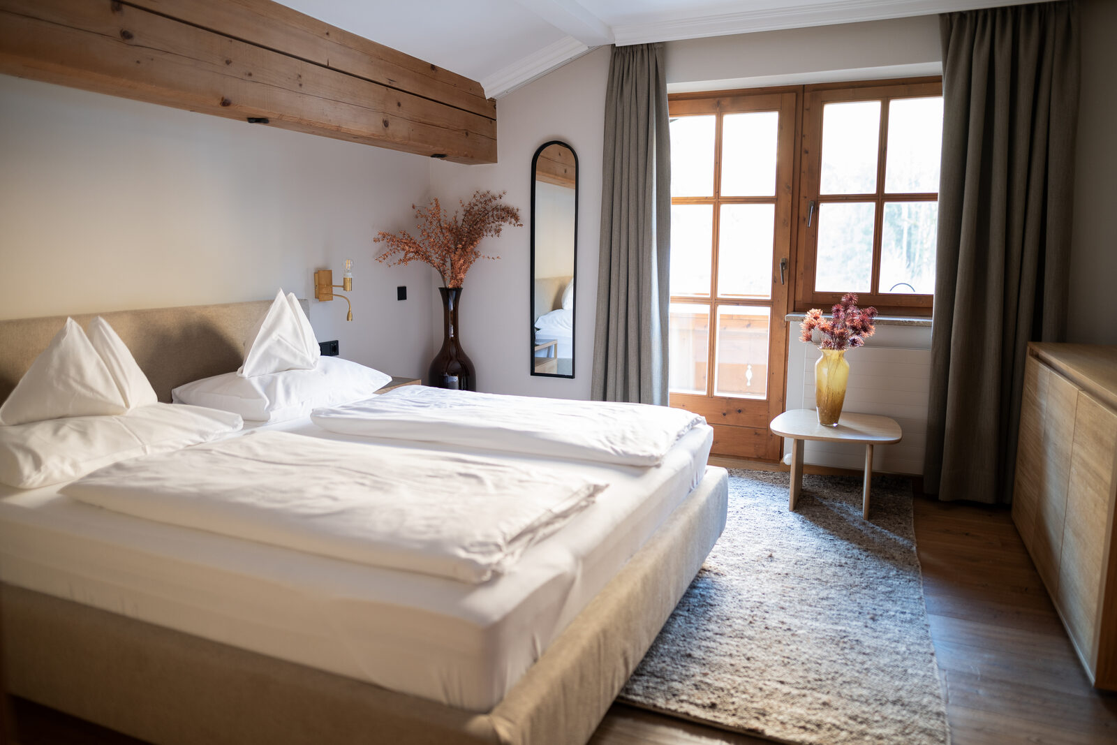 Bedroom suite with timber-beam ceiling, linen curtains and an oval mirror.