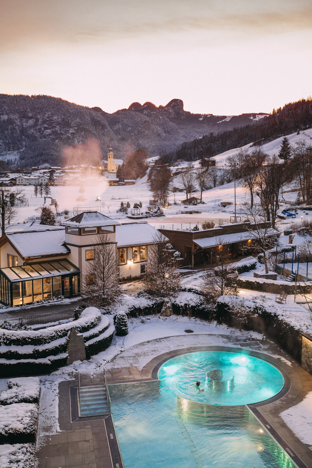POST in winter from above — snow on the grounds, the heated pool steaming, mountains behind.