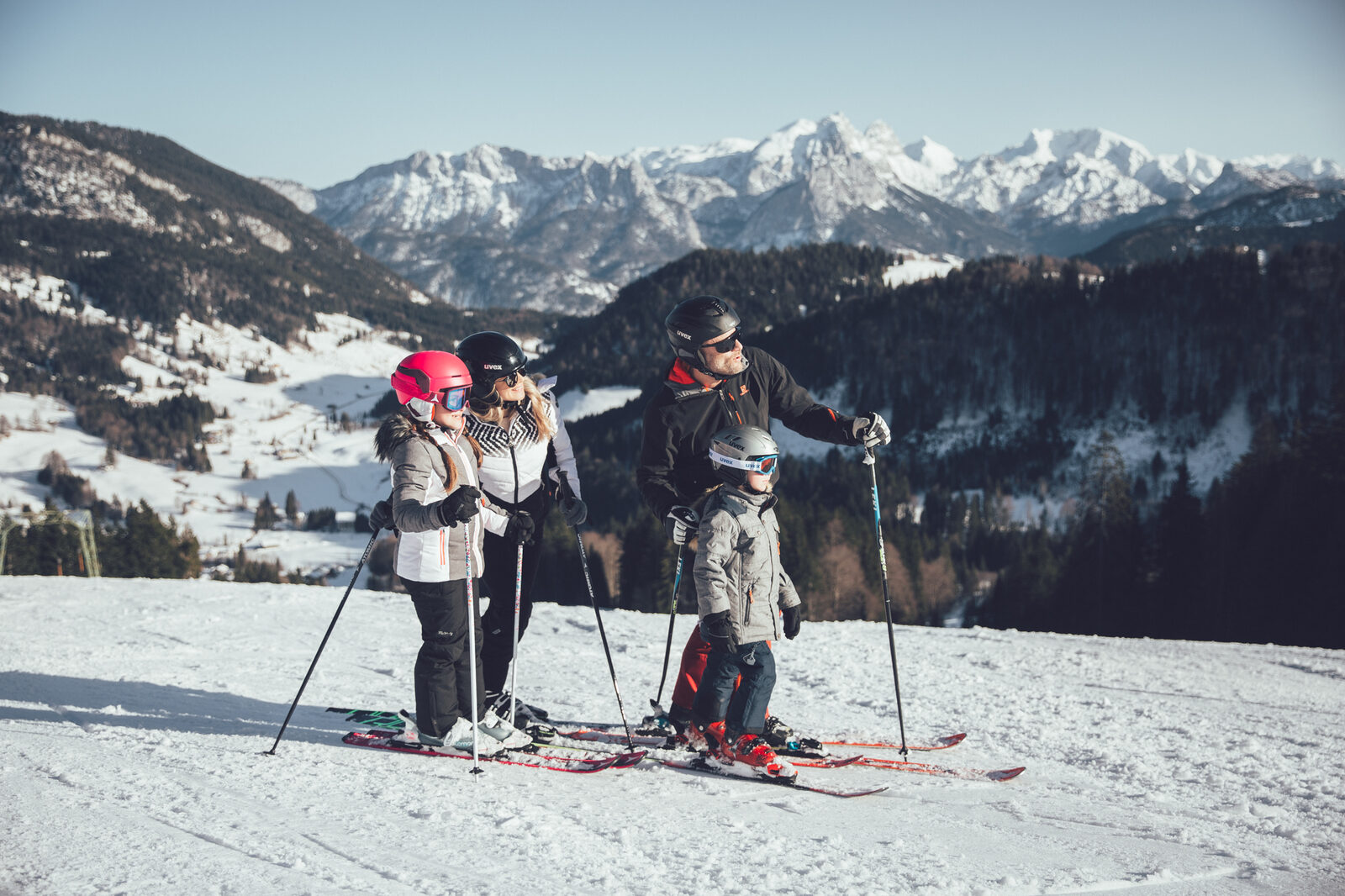 A family pauses on a sunlit piste above the valley.